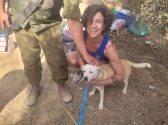 עובדת בעמותה מחייכת ורוכנת לעבר כלב עם פרווה בצבע בז' לבן ורצועה תכלת שהיא מחבקת בעוד שחייל נמצא לידם ומלטף את הכלב