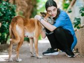 כלבה חומה לבנה, עם קולר תכלת, עומדת על מדרכה, לצידה כורעת אישה עם מכנס שחור, נעלי ספורט שחורות וחולצה כחולה קצרה, שיערה אסוף. האישה מחבקת את הכלבה ומצמידה את ראשה אל פניה.