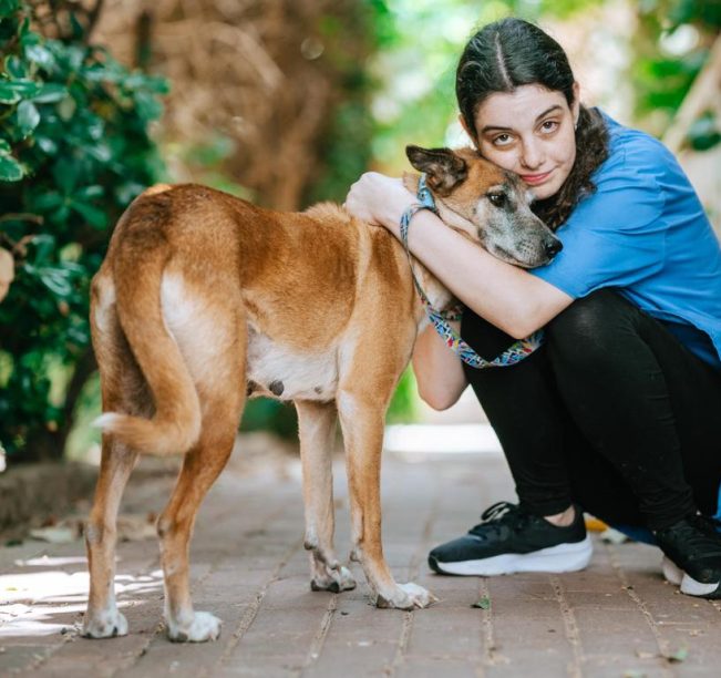 כלבה חומה לבנה, עם קולר תכלת, עומדת על מדרכה, לצידה כורעת אישה עם מכנס שחור, נעלי ספורט שחורות וחולצה כחולה קצרה, שיערה אסוף. האישה מחבקת את הכלבה ומצמידה את ראשה אל פניה.