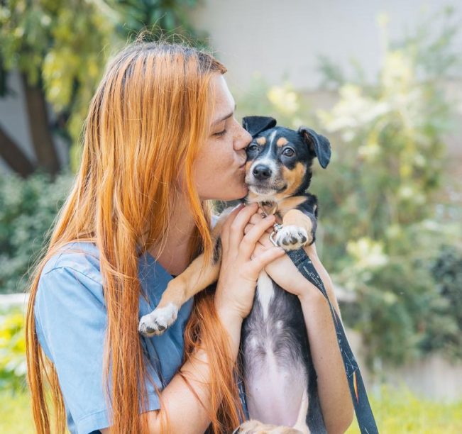 בחורה עם שיער צבוע ג'ינג'י ארוך, מכנסי ג'ינס וחולצה כחולה קצרה, מרימה גורה חומה, שחורה, לבנה ומנשקת אותה.