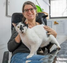 אישה עם שיער אסוף בקוקו ושקפיים, לובשת חולצה אפורה ארוכה וג'ינס ויושב על כיסא משרדי. היא מלטפת חתול שחור לבן, שעומד על רגליה.