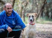 גבר, מקריח, משקפיים על פניו וזיפי שפם וזקן, לבוש סווצ'רט כחול ומכנסיים שחורות, יושב כשברכיו מכופפות, הוא מחזיק ביד אחת כלב ברצועה תכלת ובידו השנייה הוא מלטף אותו. הכלב, חום לבן בגודל בינוני, יושב לימינו, קולר אדום לצווארו, פיו פתוח והוא מפנה ראשו שמאלה. הם יושבים בחורשה. הגבר מביט על הכלב ומחייך.